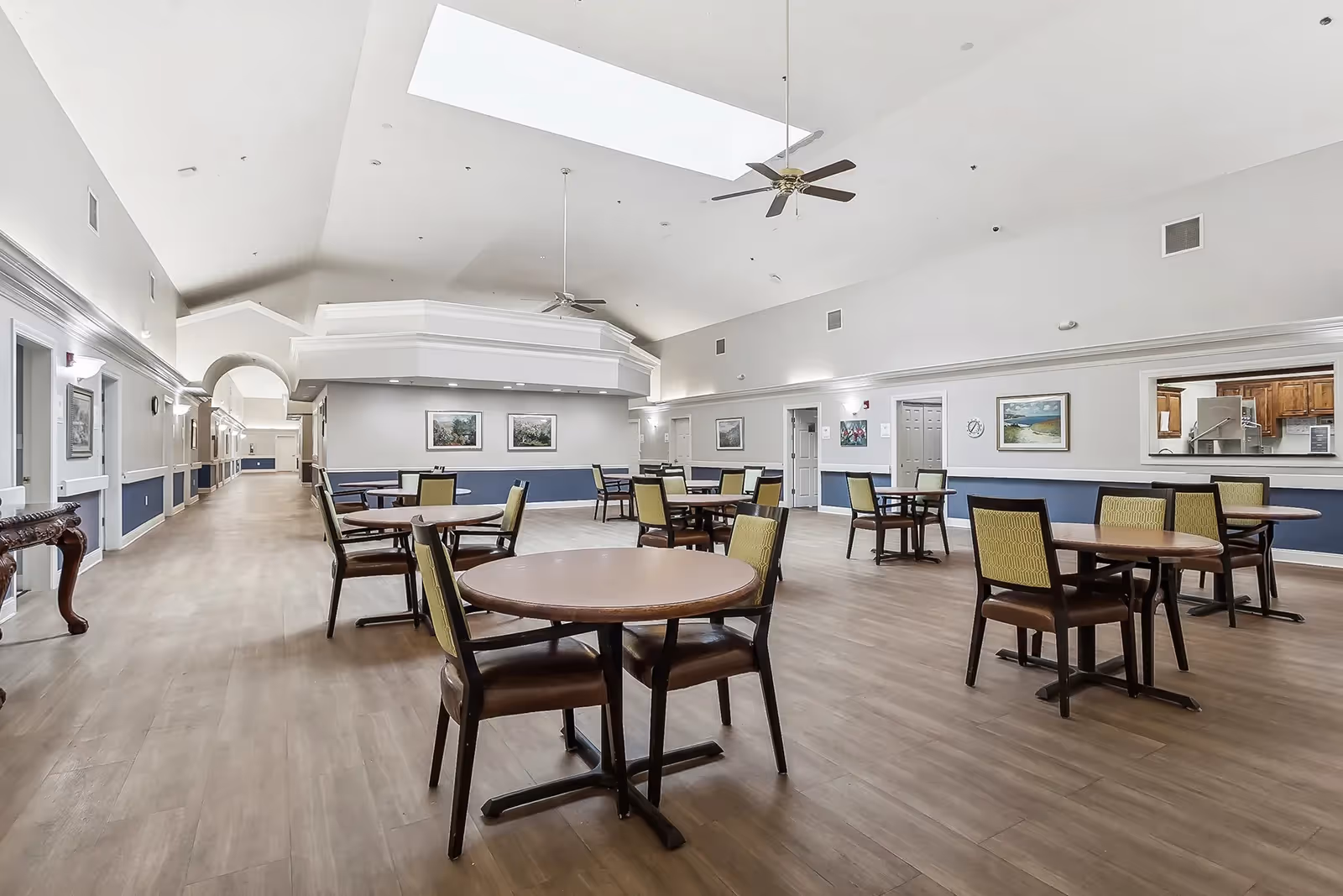 Spacious dining area with multiple round tables and chairs arranged neatly on a wooden floor. The room has high ceilings with two ceiling fans and a large skylight. Walls are painted white with blue wainscoting and decorated with framed artwork. There is a hallway leading to other rooms and a window opening to a kitchen area.