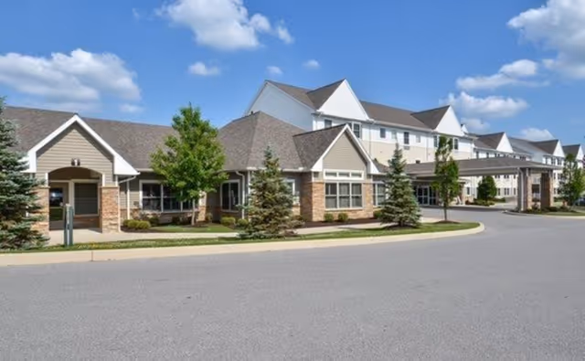 Exterior view of a senior living facility building with multiple peaked roofs, beige siding, stone accents, and several windows. The building is surrounded by small trees and shrubs, with a paved driveway in front under a partly cloudy blue sky.