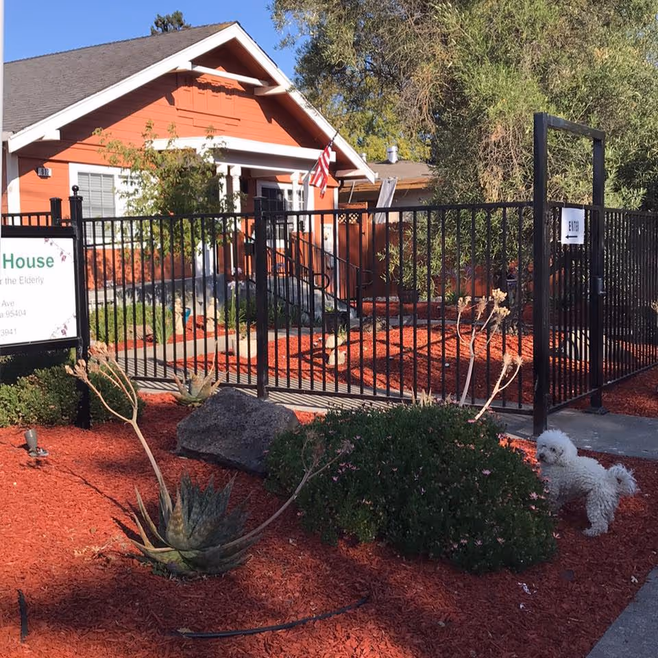 Front exterior view of a single-story orange house with white trim, surrounded by a black metal fence. The yard is landscaped with red mulch, various plants, and a small white dog standing near the sidewalk. An American flag is mounted near the entrance, and a sign partially visible reads 'Ashkalon House'.