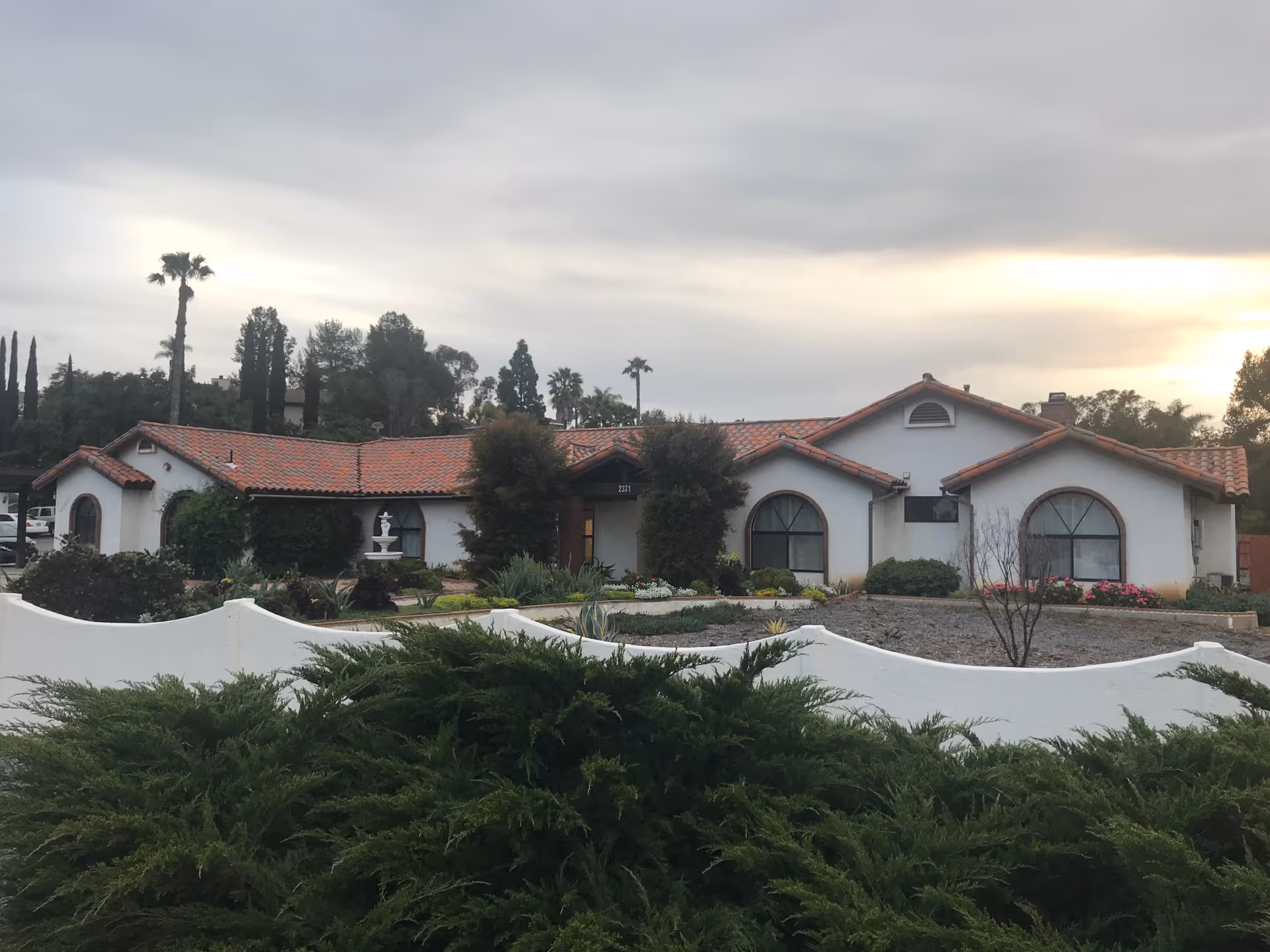 Single-story stucco building with red tile roof, arched windows and landscaped front yard behind a low white wall under an overcast sky.