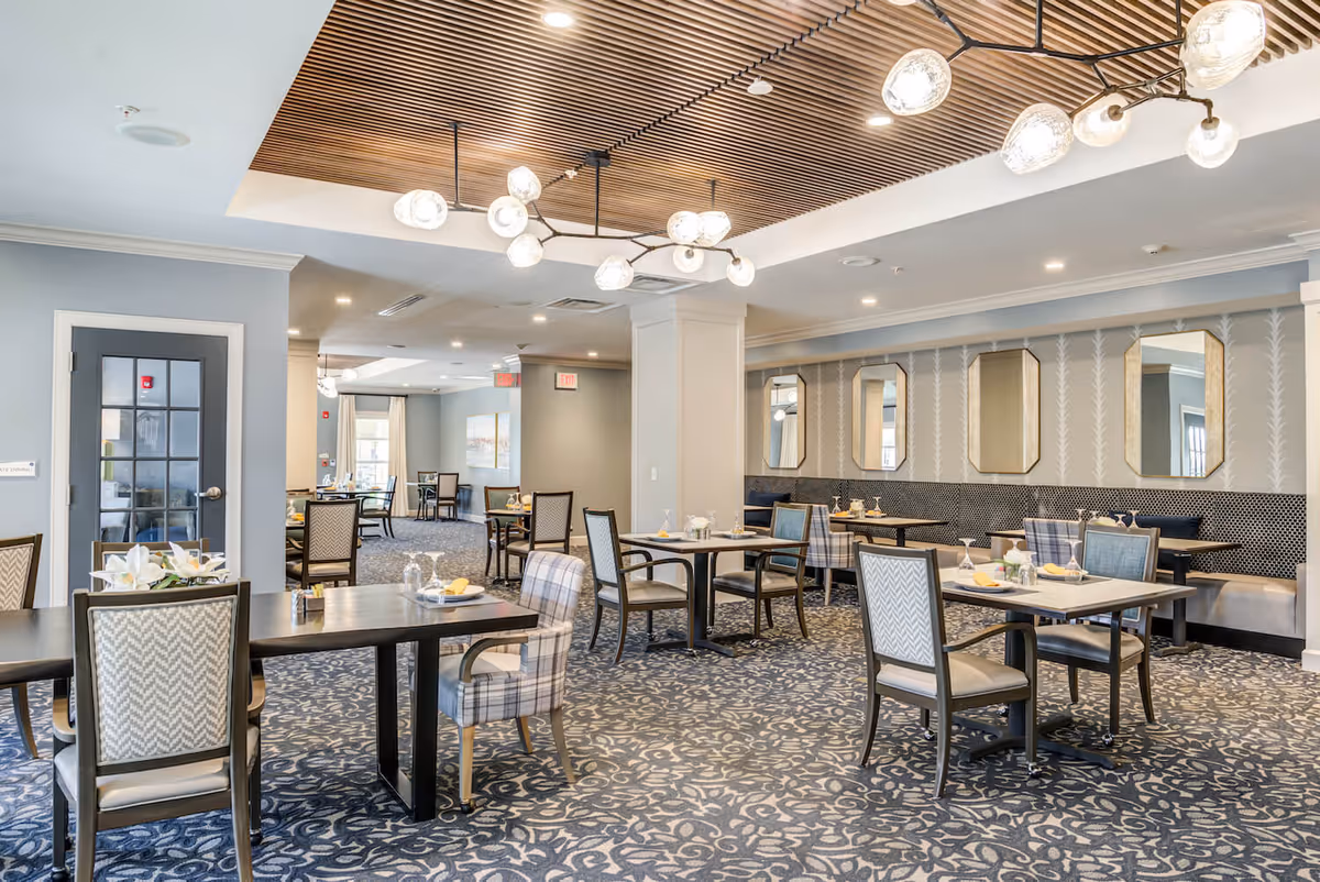 A spacious and elegant dining room with multiple tables and chairs arranged neatly. The room features a patterned carpet, a wooden slatted ceiling section with modern light fixtures, and a wall with decorative mirrors. Tables are set with plates, glasses, and napkins, creating a welcoming atmosphere.