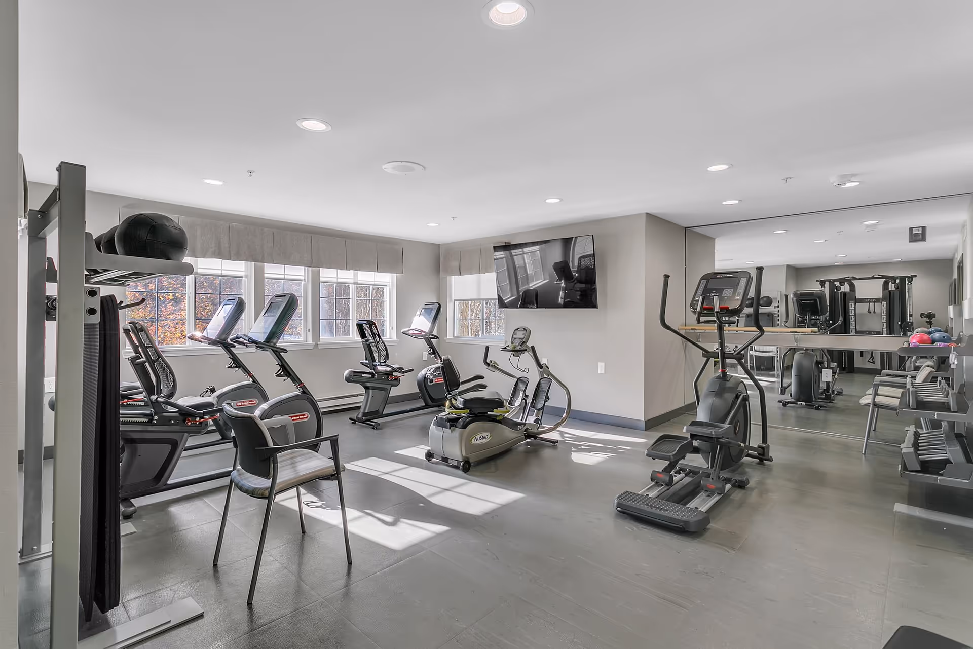 A bright fitness room with exercise equipment including stationary bikes, an elliptical machine, and weight training machines. The room has large windows letting in natural light, a wall-mounted TV, and a large mirror on one wall. There is also a chair and some exercise balls on a rack.