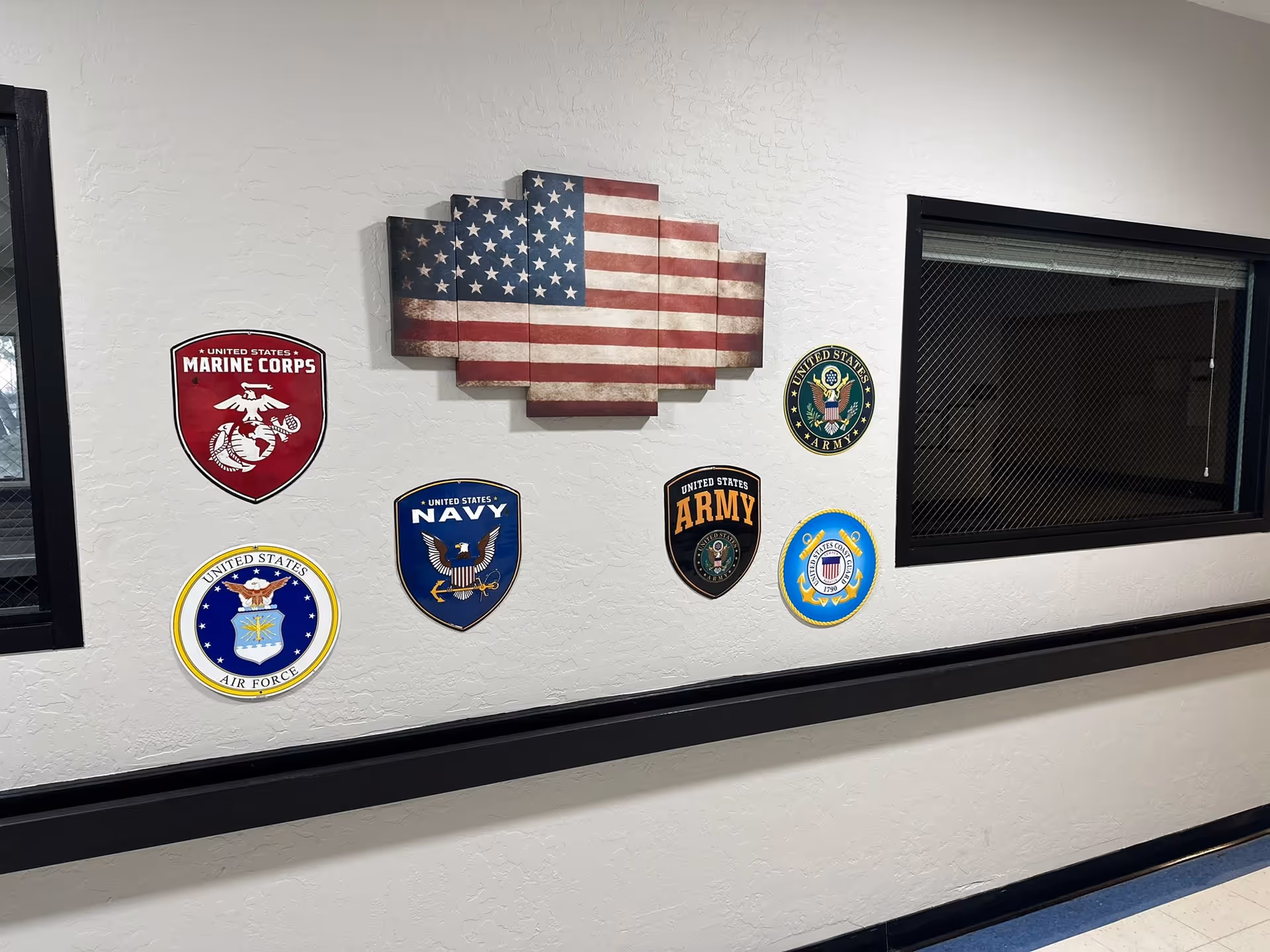 Interior hallway wall decorated with plaques of United States military branches including Marine Corps, Navy, Air Force, Army, and Coast Guard, along with a multi-panel American flag artwork. The wall is white with a textured finish and has two windows with black frames.