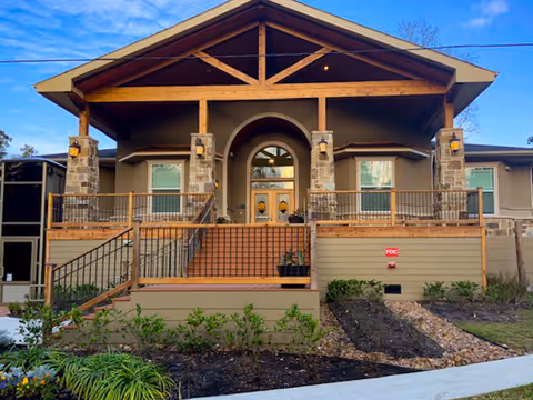 Front exterior view of a building with a wooden porch supported by stone pillars, stairs leading up to double doors with glass panels, and landscaped garden beds in front.