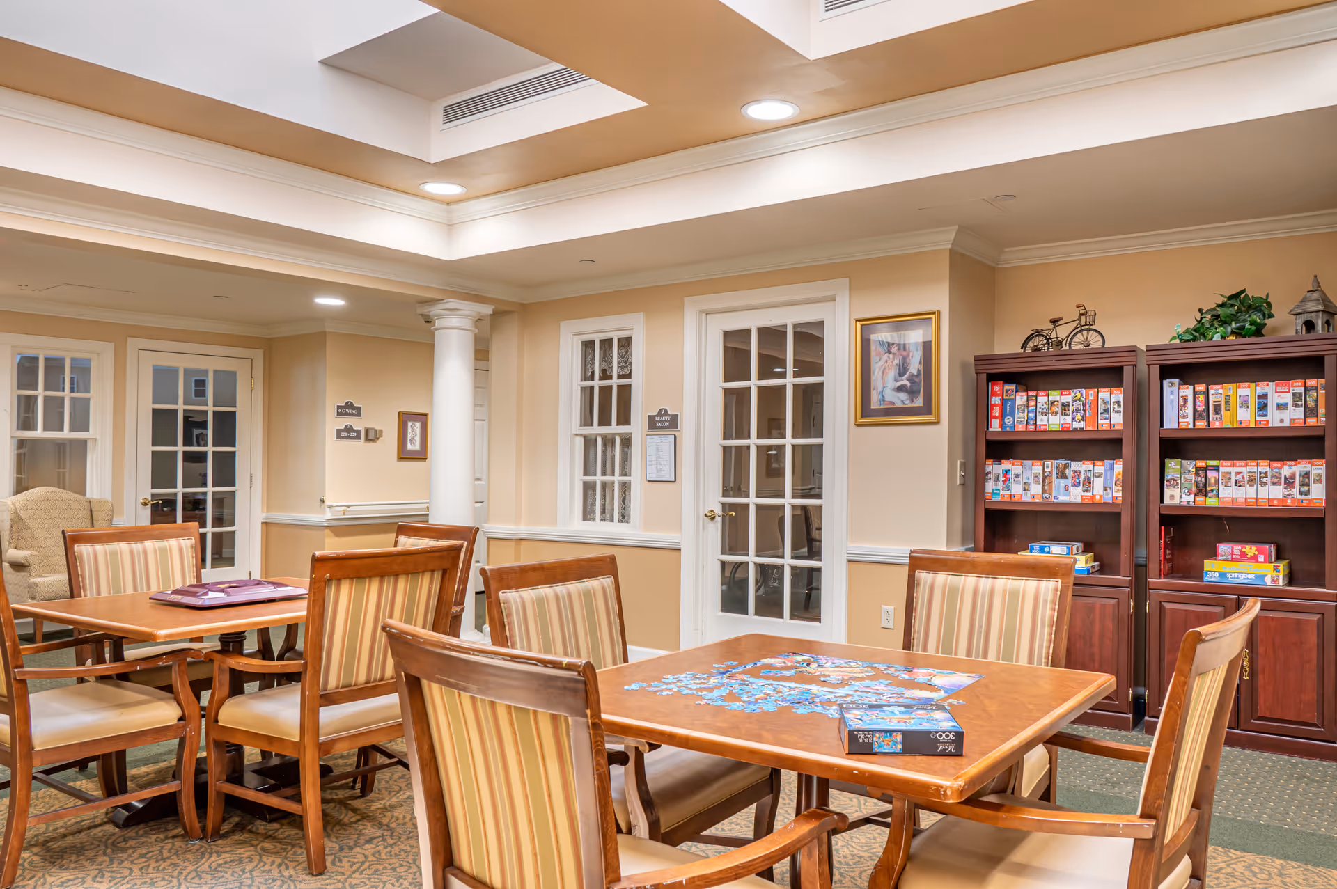 Well-lit communal activity room with wooden tables and chairs, a puzzle in progress on one table, and shelves of board games.