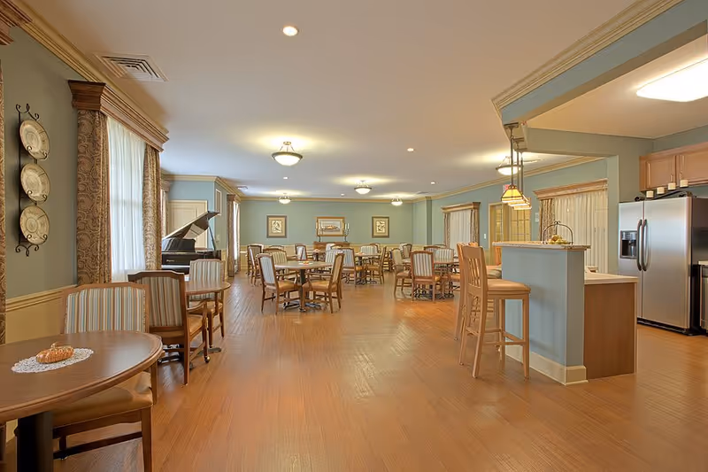 Spacious dining room with multiple round wooden tables and chairs, a grand piano near the window, soft lighting from ceiling fixtures, and a kitchen area with a breakfast bar and stainless steel refrigerator in the background.