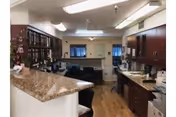 Interior view of a kitchen area with wooden cabinets, granite countertops, and various kitchen appliances. The space has a wooden floor and is well-lit with ceiling lights. There is a counter with a raised bar area and a sink visible on the right side.