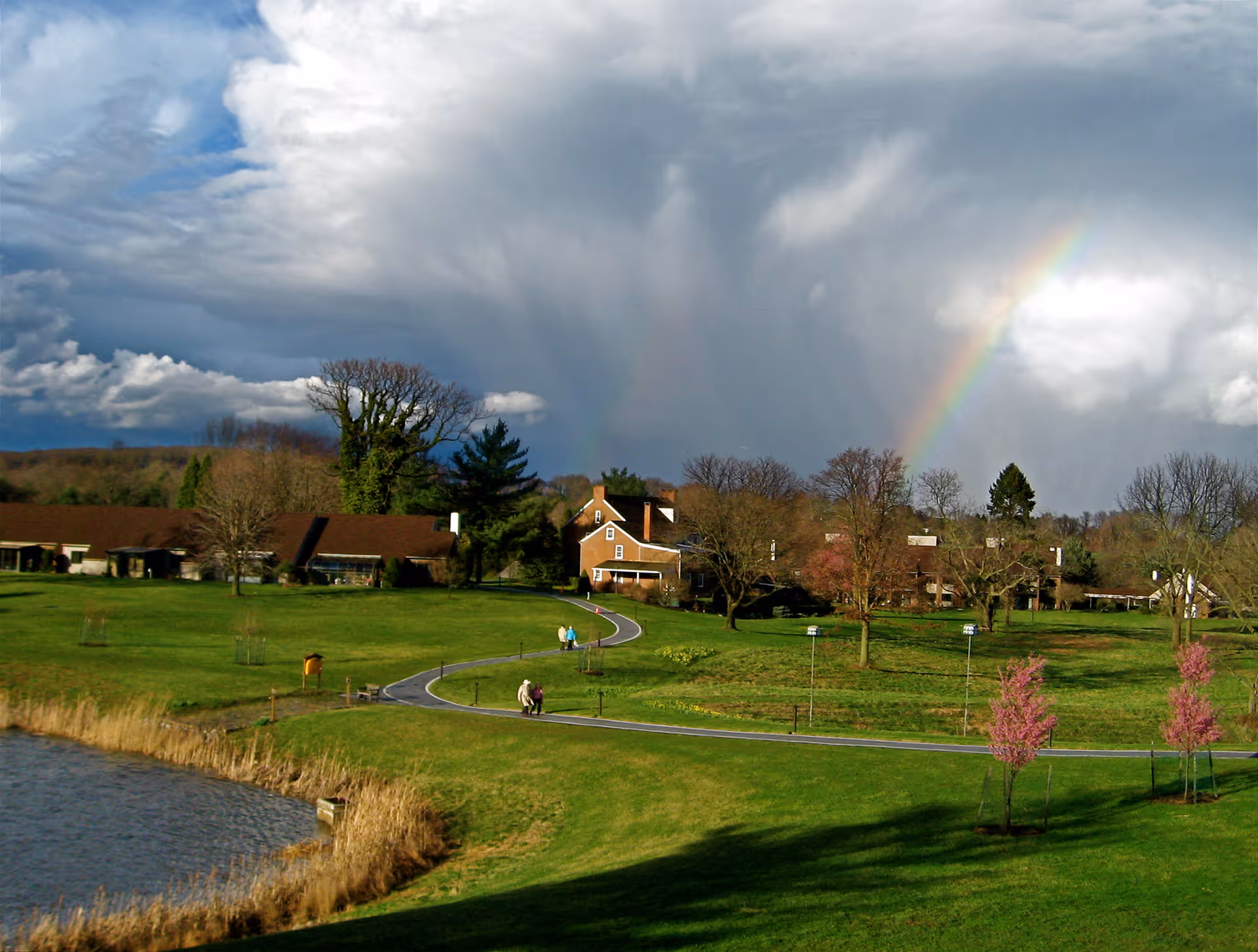 A scenic view of a senior living facility with green lawns, a winding paved path, several trees, and buildings with brown roofs. There is a body of water in the foreground and a rainbow visible in the cloudy sky above.