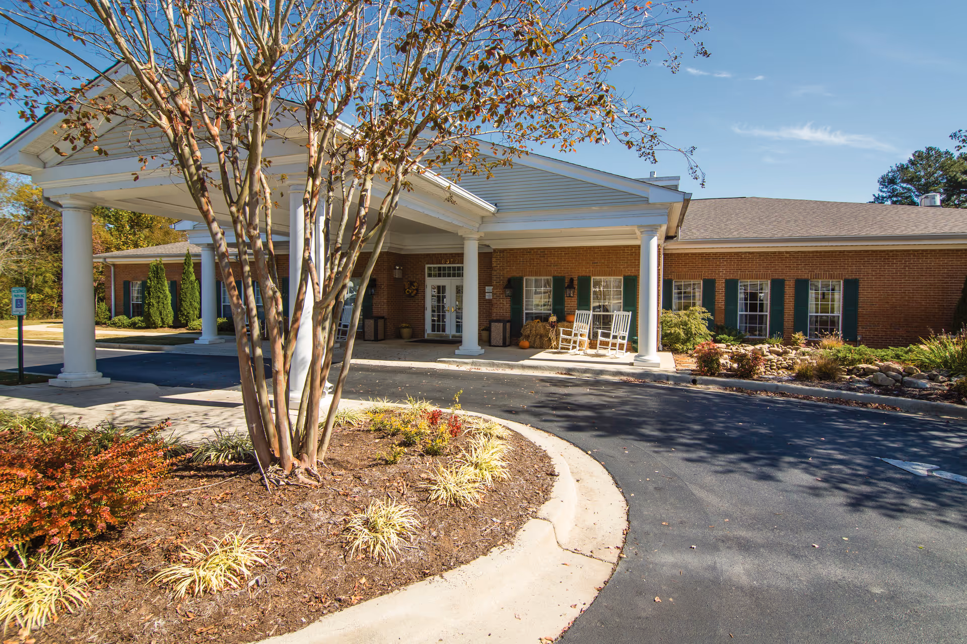 Exterior view of Brookdale Hampton Cove facility showing a brick building with green shutters, a covered entrance supported by white columns, rocking chairs on the porch, and landscaped plants and trees in front under a clear blue sky.