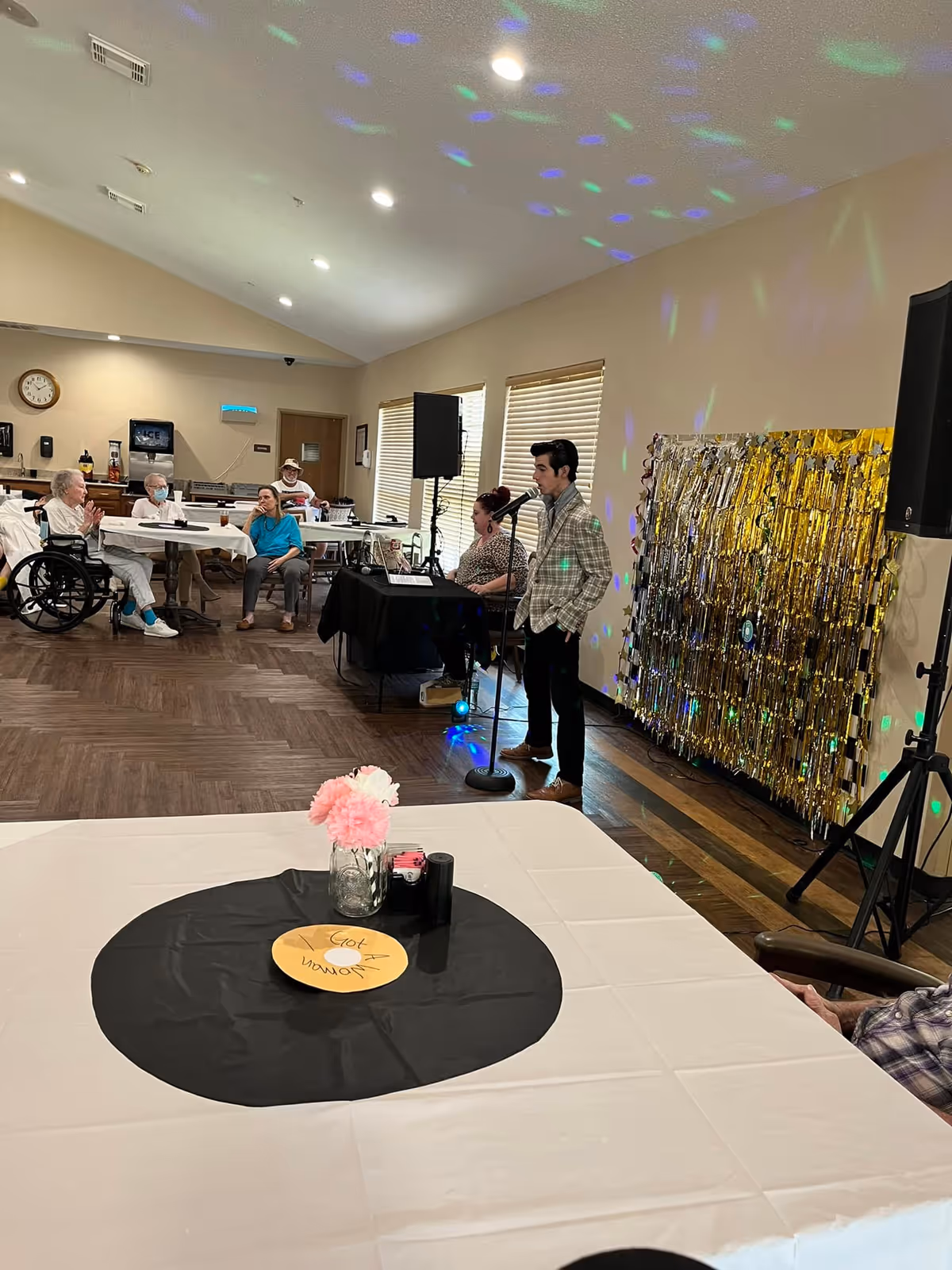 A group of elderly people seated at tables in a common room of a nursing home, watching a young man speaking into a microphone next to a woman seated at a keyboard. The room has a festive atmosphere with colorful lights projected on the walls and a gold tinsel backdrop behind the performers. A table in the foreground is decorated with a black circular cloth, a yellow paper record, and a vase with pink flowers.