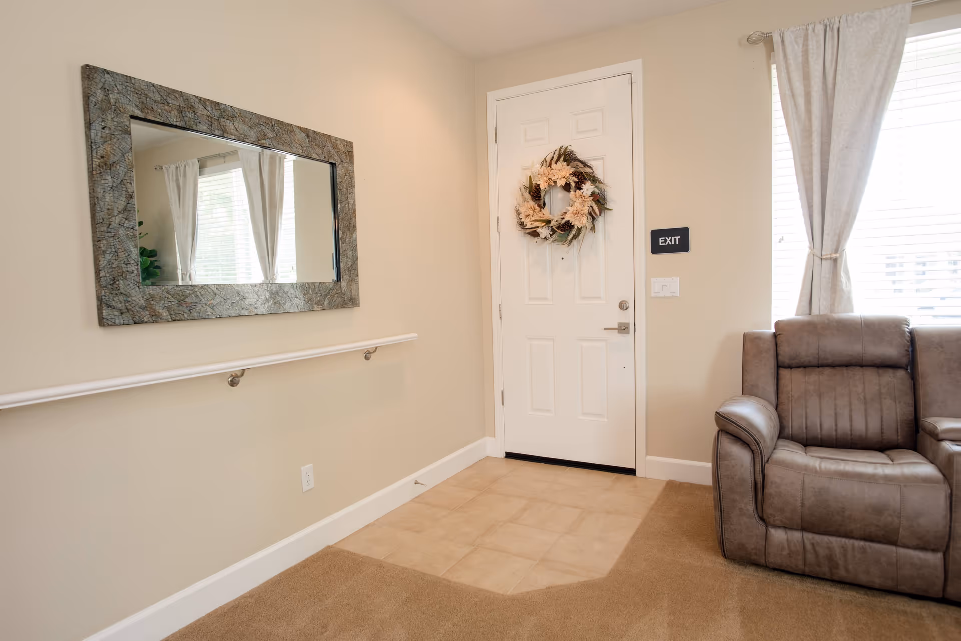 Interior view of a room with a white door decorated with a floral wreath, a large rectangular mirror with a textured frame on the wall, a handrail below the mirror, beige walls, a window with white curtains, and a gray cushioned armchair partially visible on the right.