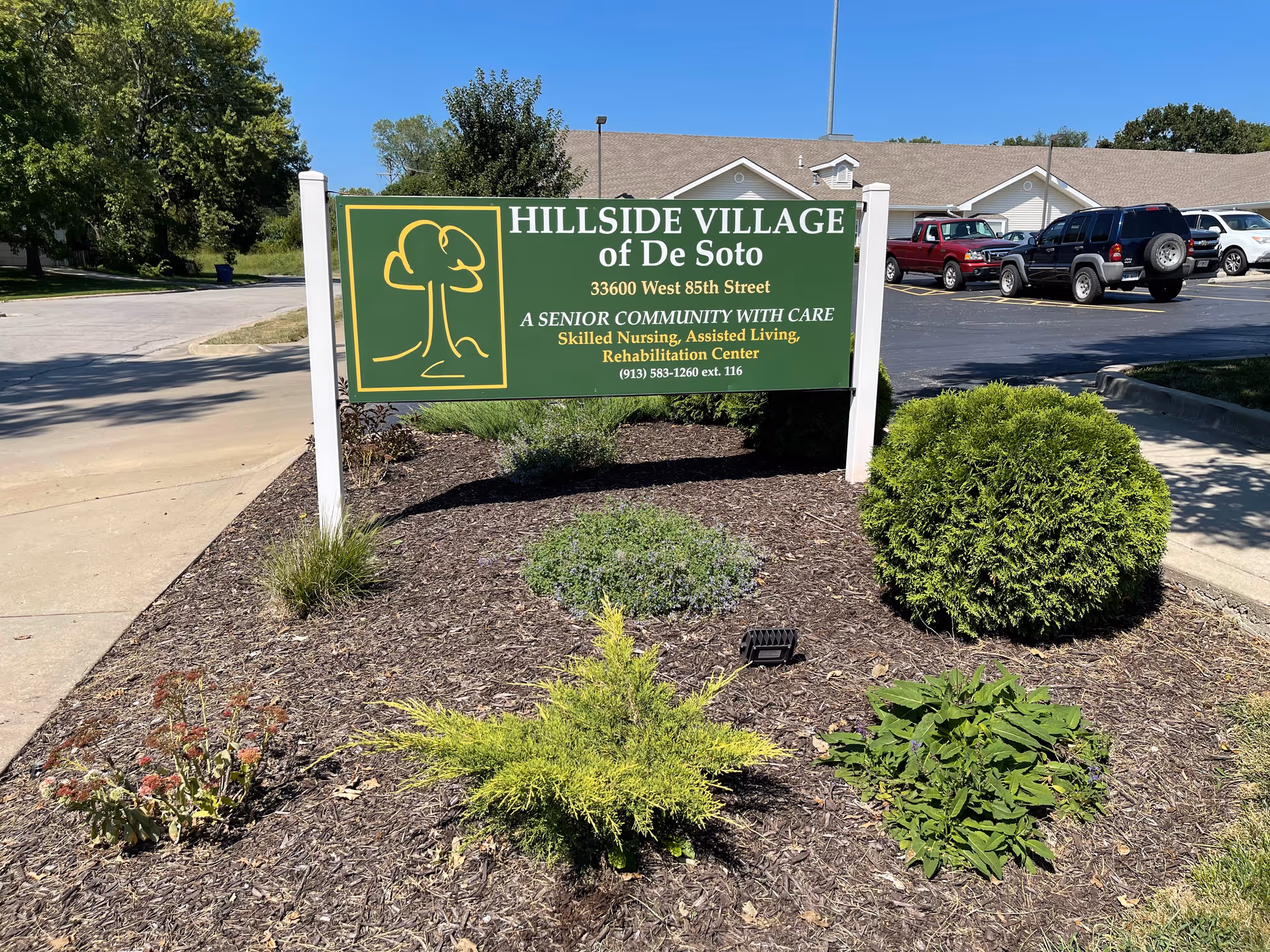 Green sign reading 'HILLSIDE VILLAGE of De Soto' in a mulch planting bed with shrubs in front of the facility and parking lot.