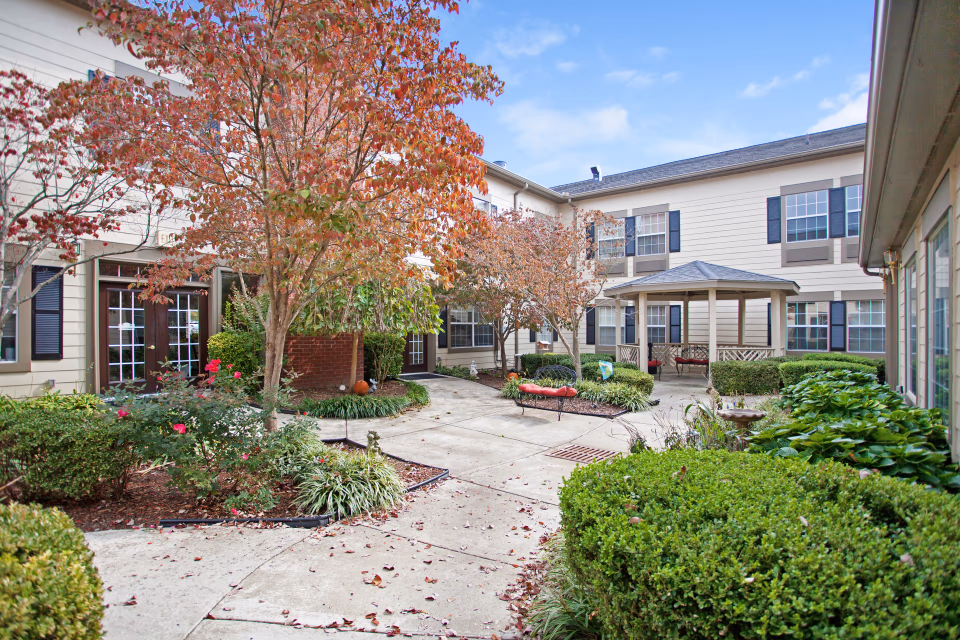 Outdoor courtyard area of The Carriages of Rivergate senior living facility featuring a paved walkway surrounded by landscaped bushes, trees with autumn foliage, and flower beds. There is a gazebo with benches and the building's beige exterior with multiple windows and black shutters is visible in the background.