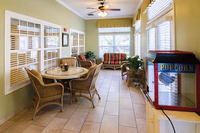 A bright indoor seating area with a round table surrounded by four wicker chairs on the left, a striped cushioned loveseat and chair at the far end near large windows, a ceiling fan above, and a popcorn machine on the right side near the window.