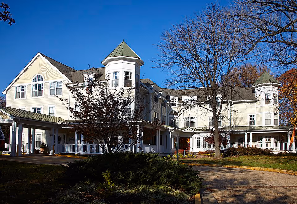 Exterior view of a large, multi-story senior living facility building with light-colored siding, green metal roof accents, and a covered entrance. There are trees with autumn foliage and a clear blue sky in the background.