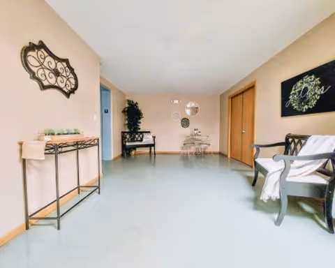 A bright and clean interior hallway or common area with light-colored walls and floor. There is a decorative metal wall hanging on the left wall above a narrow table with candles. A bench with cushions is placed against the far wall near a potted plant. On the right side, there is a wooden chair with a white blanket draped over it and a framed artwork on the wall above. A small round table with two chairs is positioned near the back wall, which is decorated with three circular mirrors or wall accents.