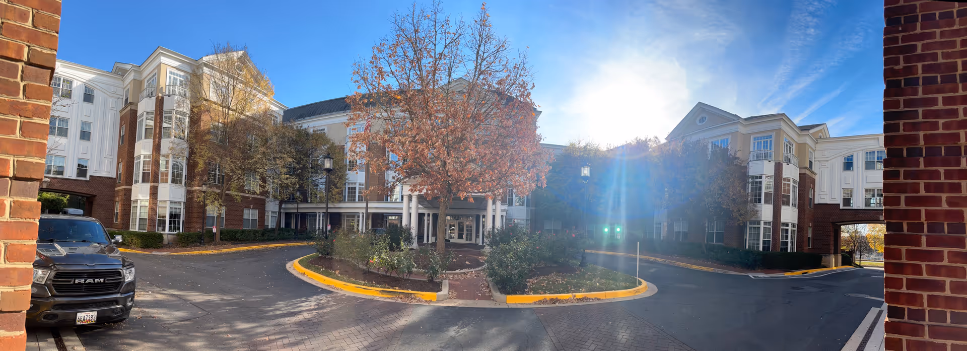 Exterior view of Grand Oaks Assisted Living Community building on a sunny day with a clear blue sky. The building features a combination of red brick and white siding with multiple windows. There is a circular driveway with a landscaped island in the center containing a tree with autumn-colored leaves and some shrubs. A black RAM truck is parked on the left side near the building.