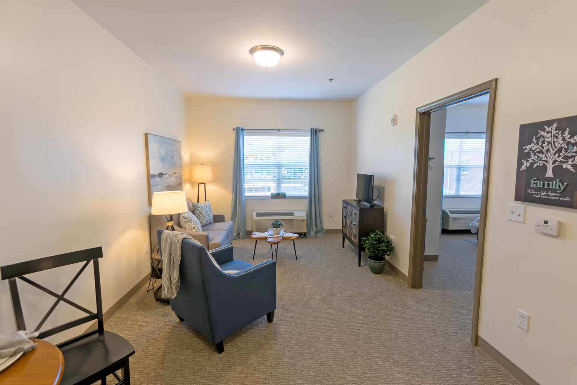 A cozy living room with a blue armchair, a gray sofa with patterned pillows, two lamps, a wooden coffee table with a small plant, a TV on a dark wooden stand, and a window with blue curtains letting in natural light. A doorway leads to a bedroom with a bed and window visible. The walls are light-colored and there is a decorative family-themed wall art near the doorway.