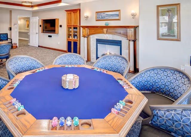 A cozy room with a large octagonal poker table covered with a blue felt surface and poker chips arranged around it. Surrounding the table are cushioned chairs with a blue circular pattern. In the background, there is a fireplace with a wooden mantle, a wall-mounted TV, and framed artwork on the walls. The room has a carpeted floor and soft lighting.