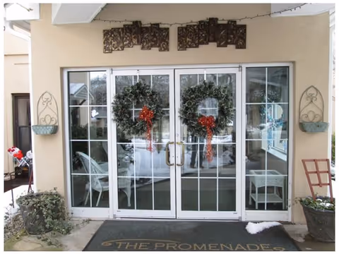 Glass double doors decorated with two large green wreaths adorned with red bows. The doors are part of a beige building entrance with potted plants on either side and a welcome mat that reads 'THE PROMENADE'. There is snow on the ground outside and some outdoor furniture visible through the glass.