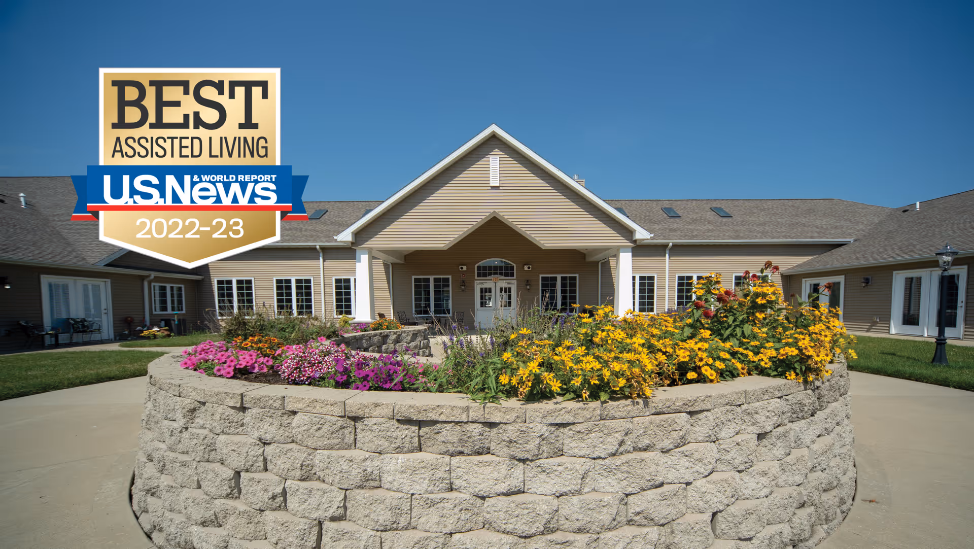 Front entrance of a single-story senior living building with a circular stone flower bed full of yellow and pink flowers under a clear blue sky.