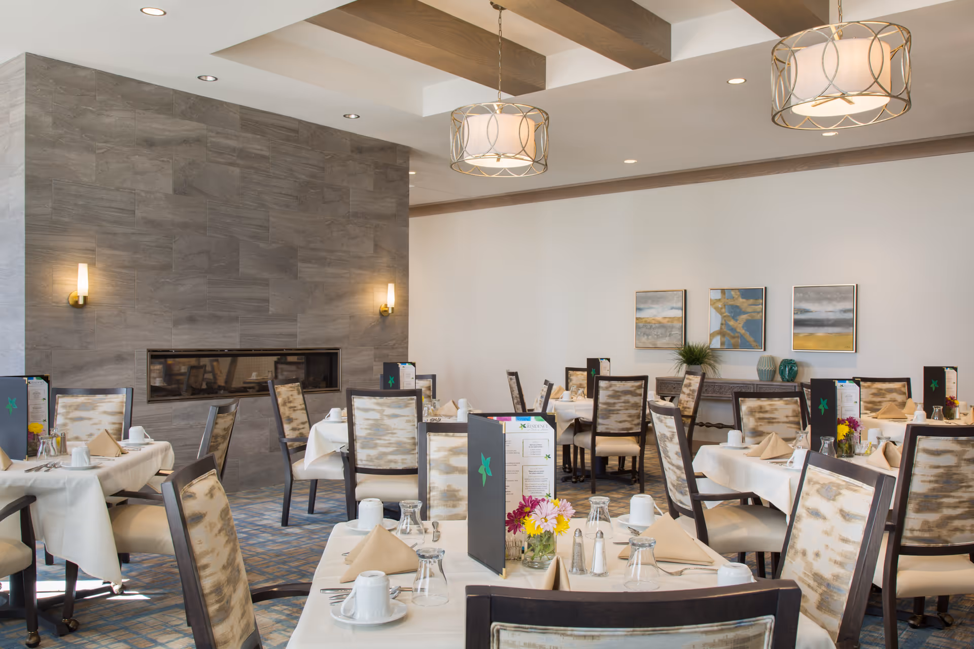 A bright and elegant dining room with multiple tables covered in white tablecloths, each set with beige napkins, white cups, glasses, and silverware. The chairs have patterned upholstery with dark wooden frames. The room features a modern gray tiled fireplace on one wall, three abstract paintings on a white wall, and two decorative pendant lights hanging from a ceiling with wooden beams. Small flower arrangements and menus are placed on the tables.