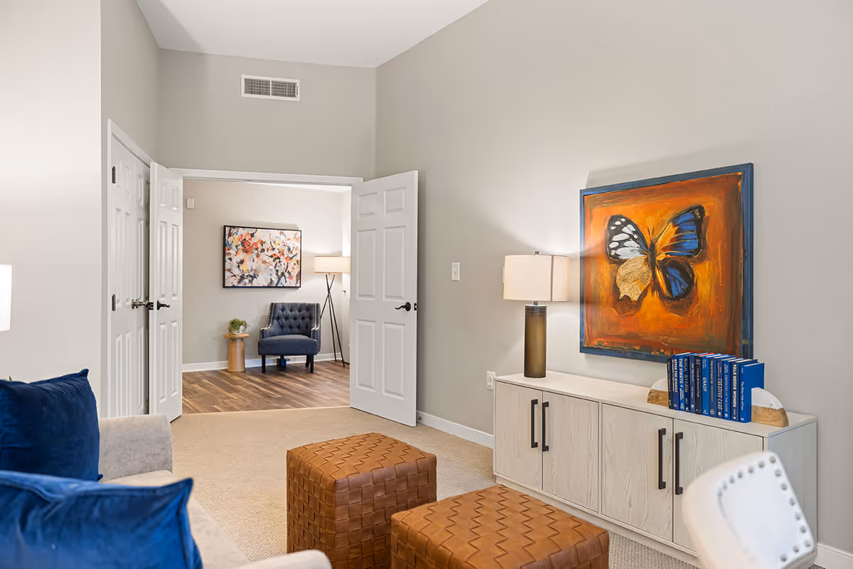 A cozy living room area with a beige sofa adorned with blue pillows, two brown woven ottomans, and a white cabinet topped with a lamp and a set of blue books. A vibrant painting of a butterfly with orange and blue colors hangs above the cabinet. In the background, an open double door reveals another room with a blue armchair, a small wooden side table with a plant, a floor lamp, and a colorful abstract painting on the wall.