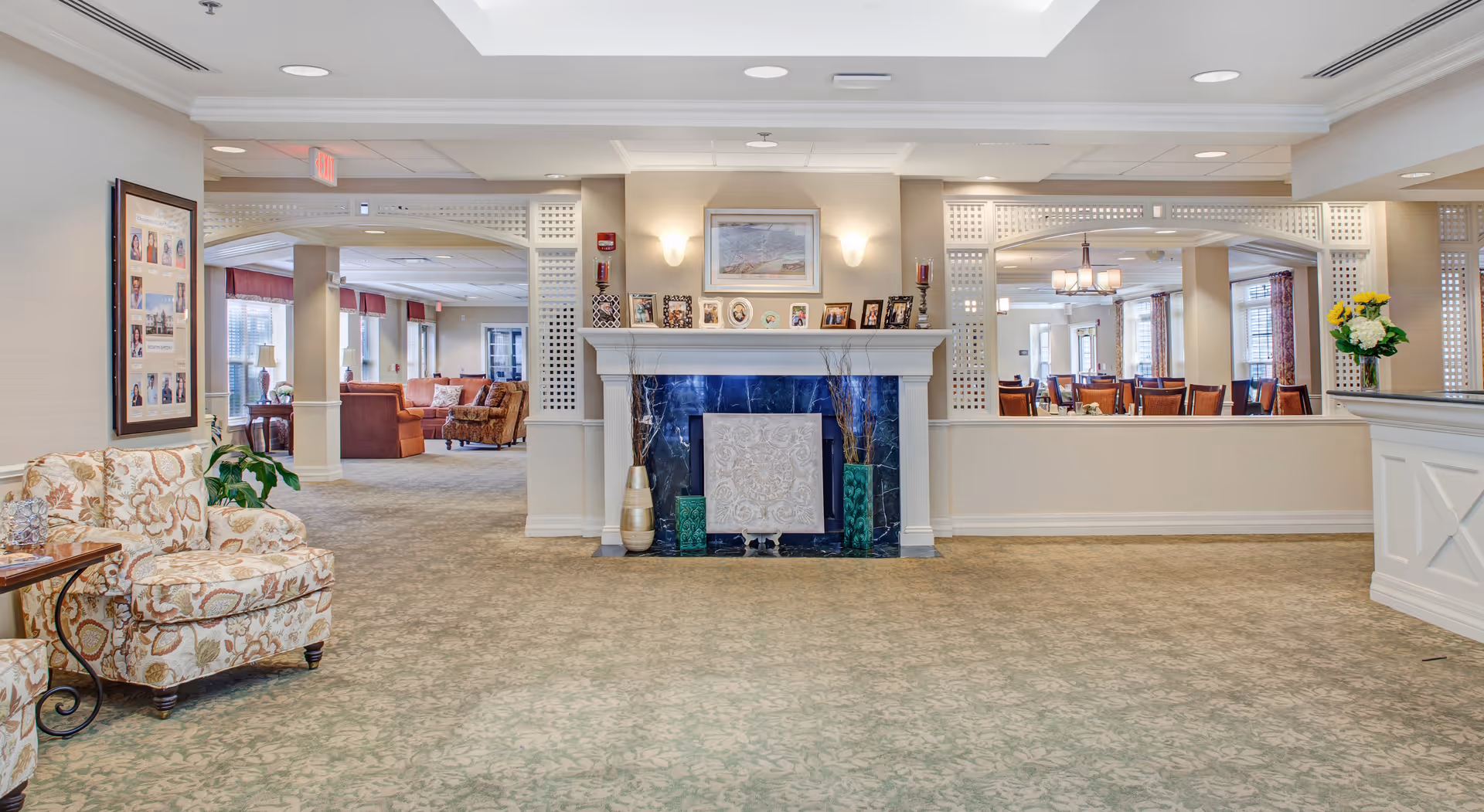 A spacious, well-lit common area in a senior living facility featuring a decorative fireplace with framed photos and art above it. To the left, there is a patterned armchair and a small side table with a plant. In the background, there are additional seating areas with sofas and chairs, and to the right, a dining area with tables and chairs is visible through a large open partition. The room has a soft green patterned carpet and neutral-colored walls.