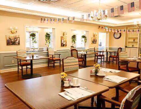 A well-lit dining room with several wooden tables and chairs arranged neatly. The tables are set with placemats, cups, and small flower vases. The room has windows with hanging plants, patriotic decorations including small American flags hanging from the ceiling, and framed pictures on the walls. There is a chandelier and a clock on the wall, with built-in shelves in the corner.