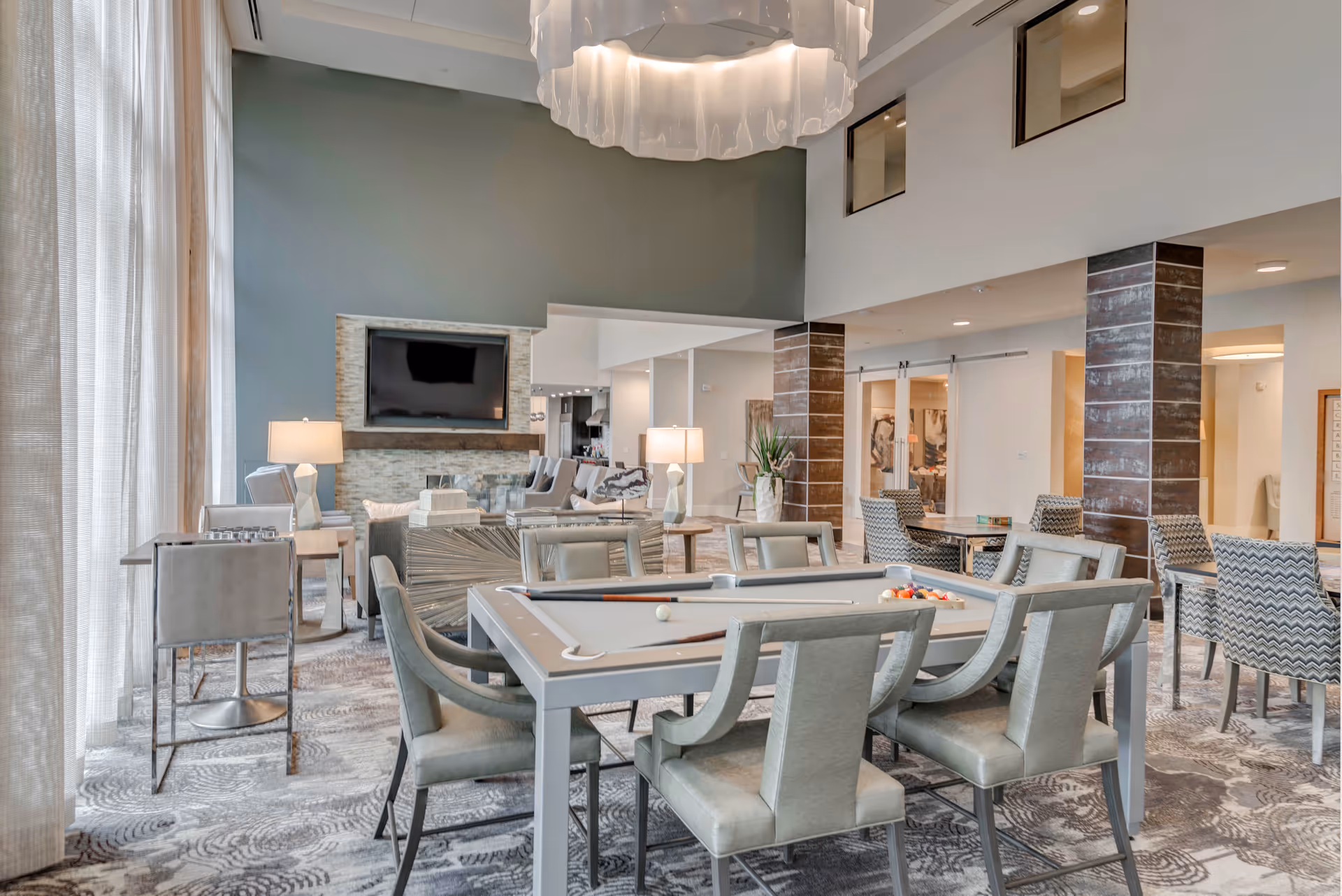 A spacious and modern common area with a pool table surrounded by chairs, a seating area with sofas and armchairs, a large flat-screen TV mounted above a stone fireplace, and large windows with sheer curtains allowing natural light to fill the room.