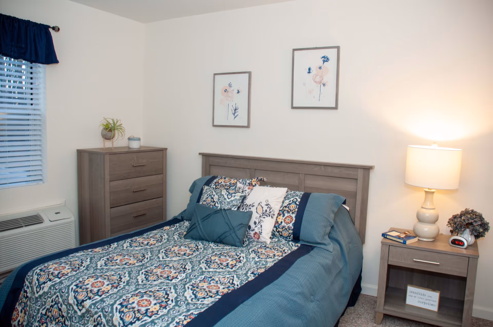 A bedroom with a bed covered in a blue patterned comforter and multiple pillows. There is a wooden headboard, a nightstand with a lamp, a clock, a book, and a small plant. On the wall above the bed are two framed floral artworks. A wooden dresser with a plant and a candle sits next to a window with blinds and a blue valance.