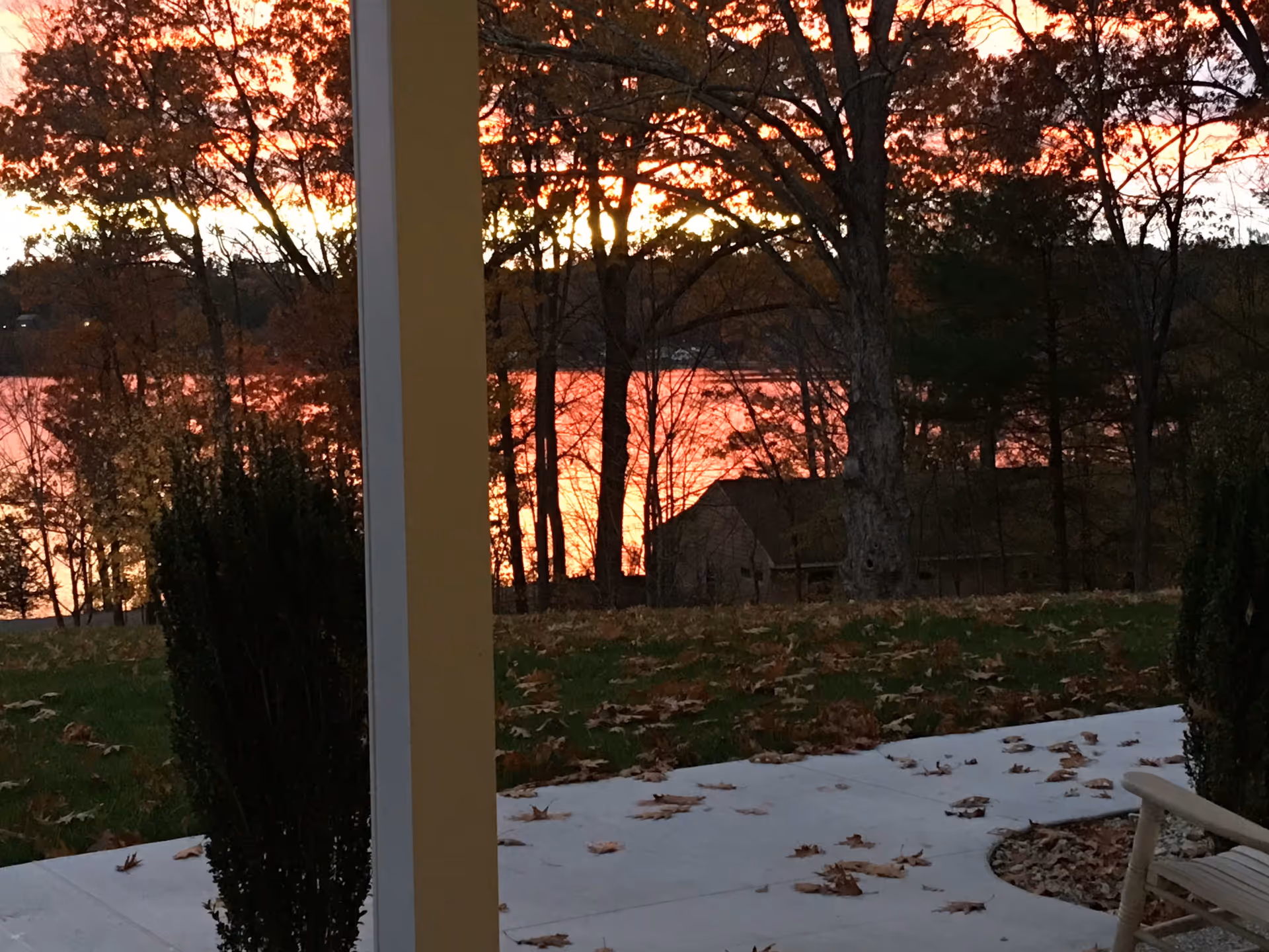 View from a porch showing a concrete walkway covered with fallen autumn leaves, a wooden chair on the right, bushes, trees with fall foliage, and a lake reflecting the orange and pink colors of a sunset in the background.