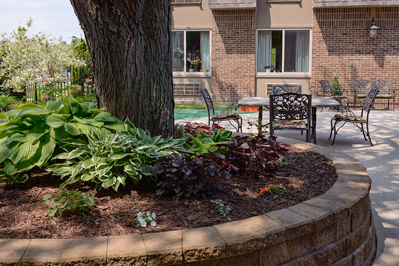 Outdoor patio area at Rivergate Terrace featuring a large tree surrounded by a raised circular brick planter filled with various green and purple plants. Several metal chairs and tables are arranged on the concrete patio near a brick building with windows.