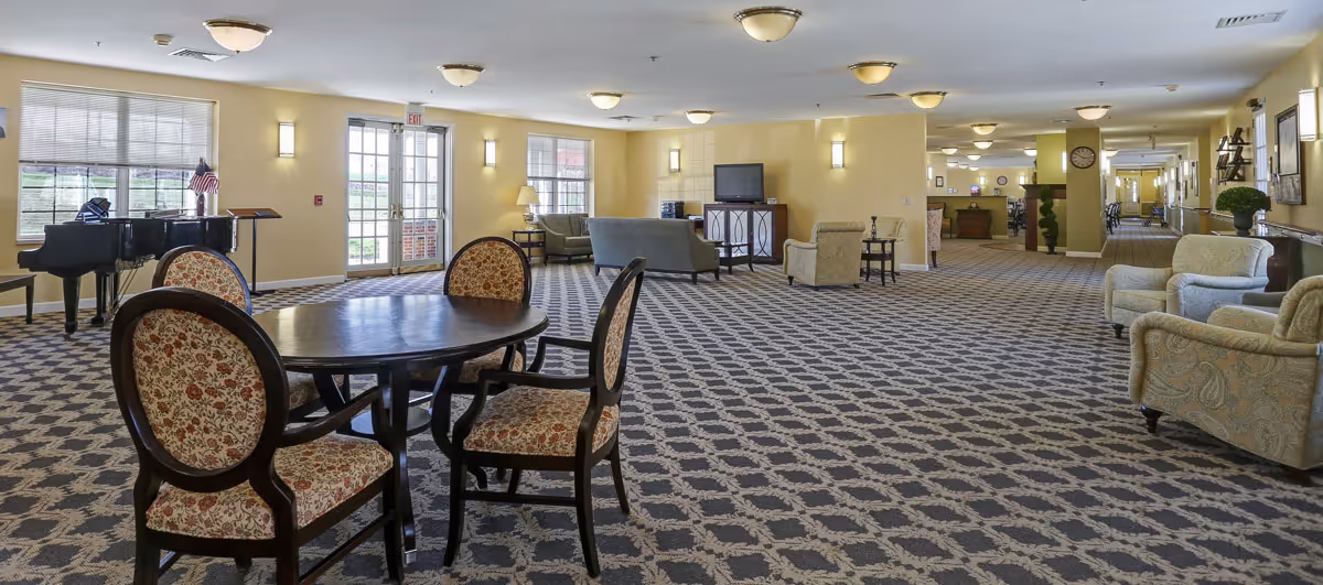 Spacious senior living facility common area with patterned carpet, round wooden table with four upholstered chairs, several armchairs and sofas, a piano near windows, and a TV on a cabinet. The room is well-lit with ceiling lights and wall sconces, and has large windows and glass doors leading outside.