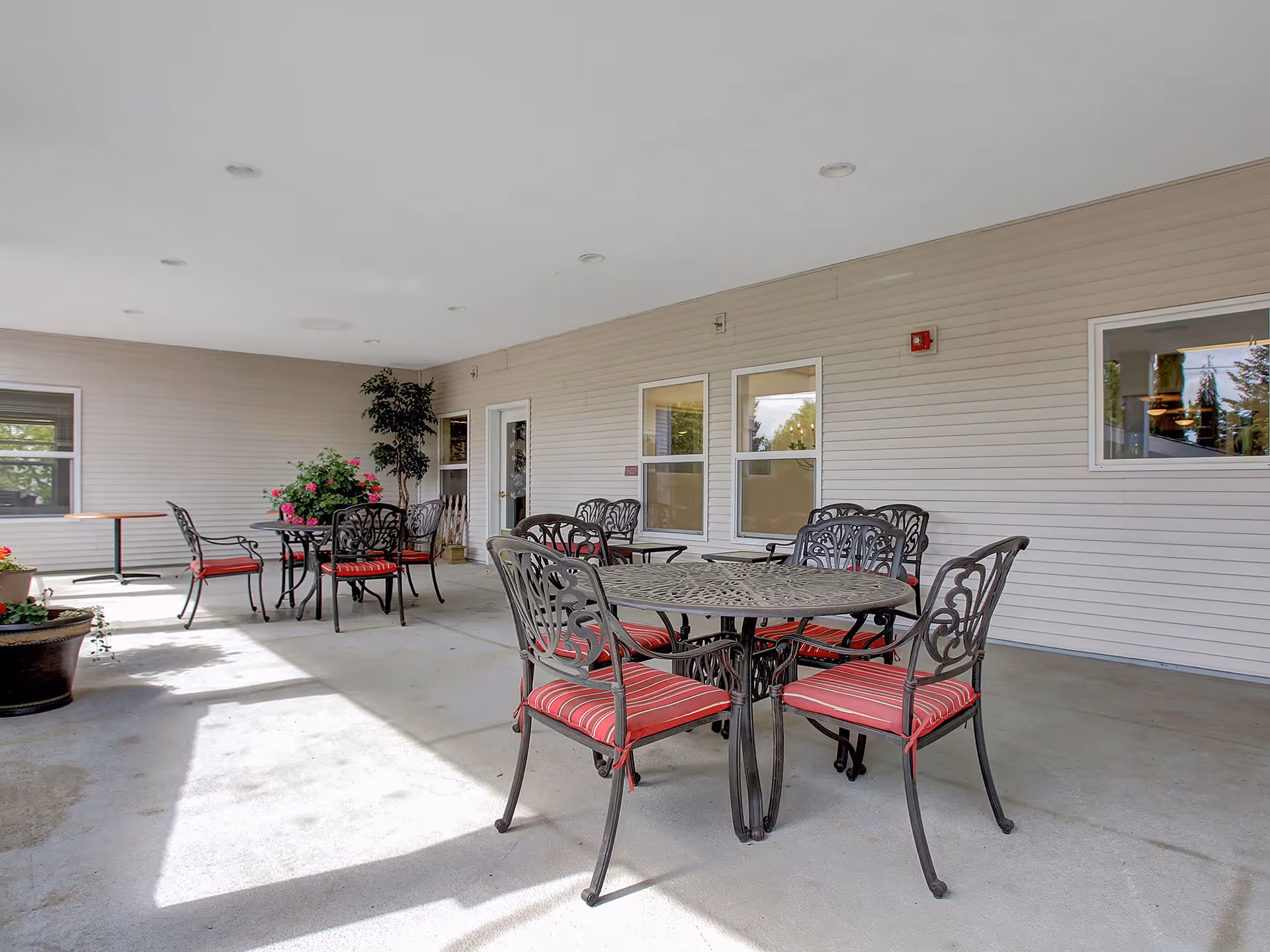 Covered outdoor patio area with several black metal tables and chairs with red striped cushions. There are potted plants and flowers near the walls, and windows and doors are visible on the beige siding wall.