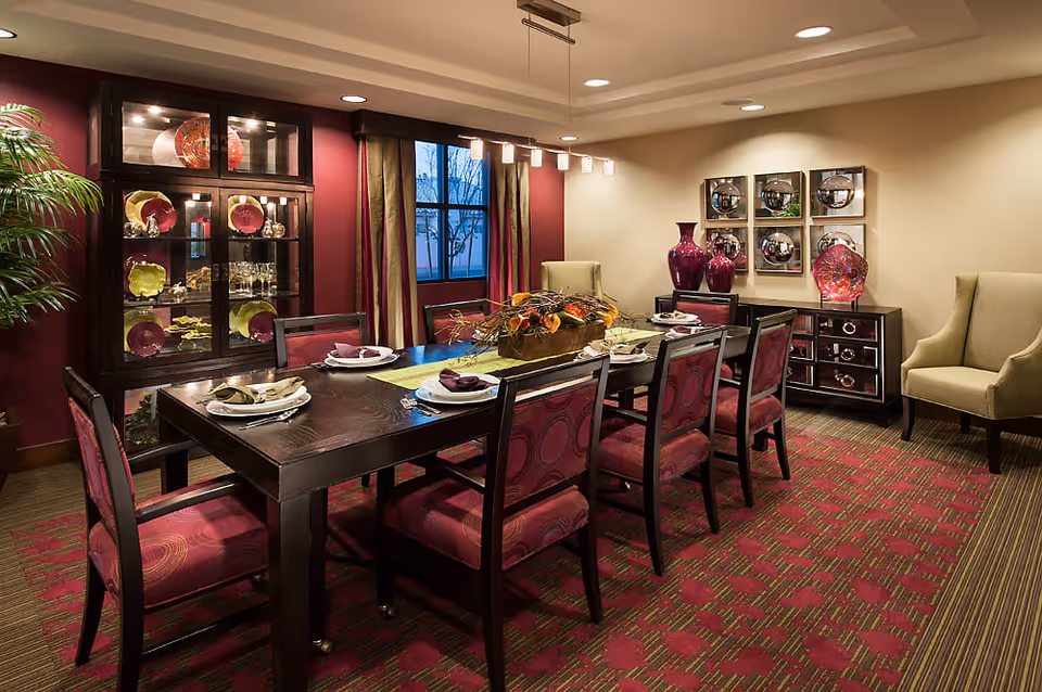 A dining room with a dark wooden table set for six, featuring purple cushioned chairs. The room has a patterned red and green carpet, a glass cabinet displaying decorative plates and glassware, and two beige armchairs. The walls are decorated with purple vases and reflective square wall art, with a window covered by curtains.