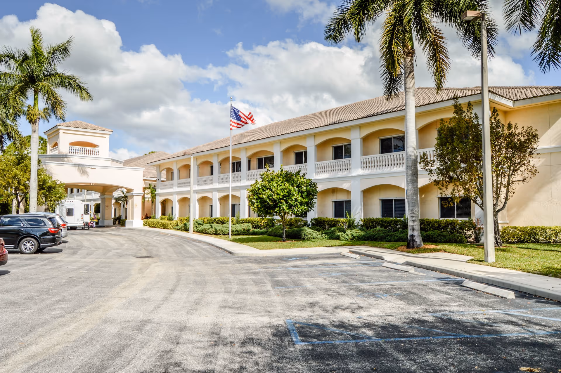 Exterior view of Courtyard Gardens of Jupiter, a two-story building with beige walls and a tiled roof, surrounded by palm trees and greenery. There is a driveway with parked cars and an American flag flying on a flagpole near the entrance under a covered portico.