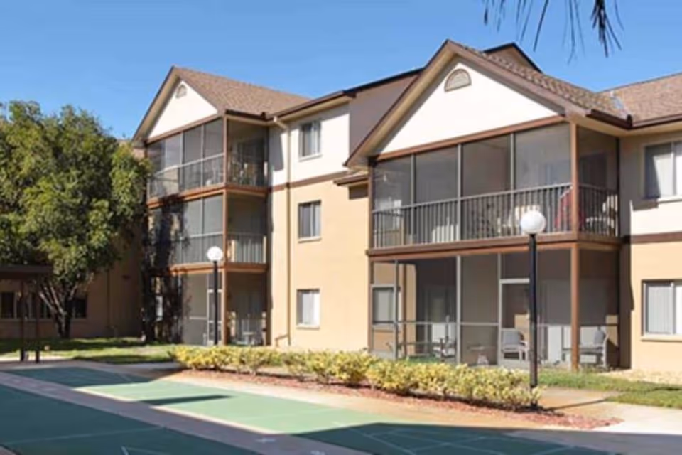Exterior view of a multi-story residential building with screened balconies and a shuffleboard court in the foreground, surrounded by greenery and trees under a clear blue sky.