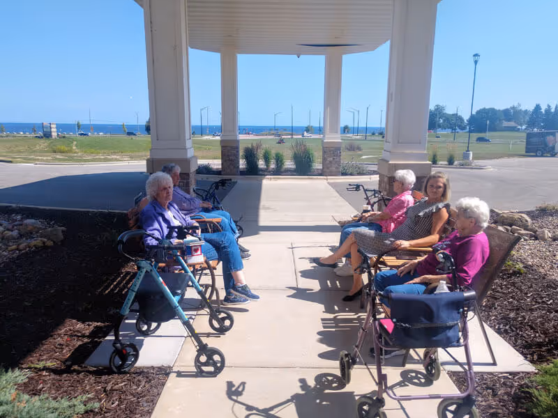 Five elderly women sitting on benches under a covered outdoor area with walkers nearby. In the background, there is a grassy area, a road, and a view of a large body of water under a clear blue sky.