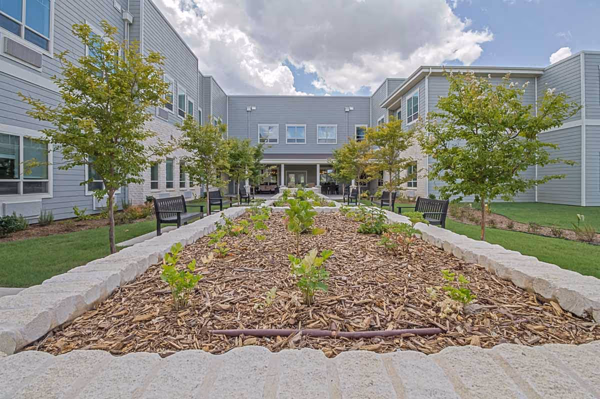 Courtyard garden with young plants, benches and trees between two wings of a gray assisted living building under a cloudy sky.