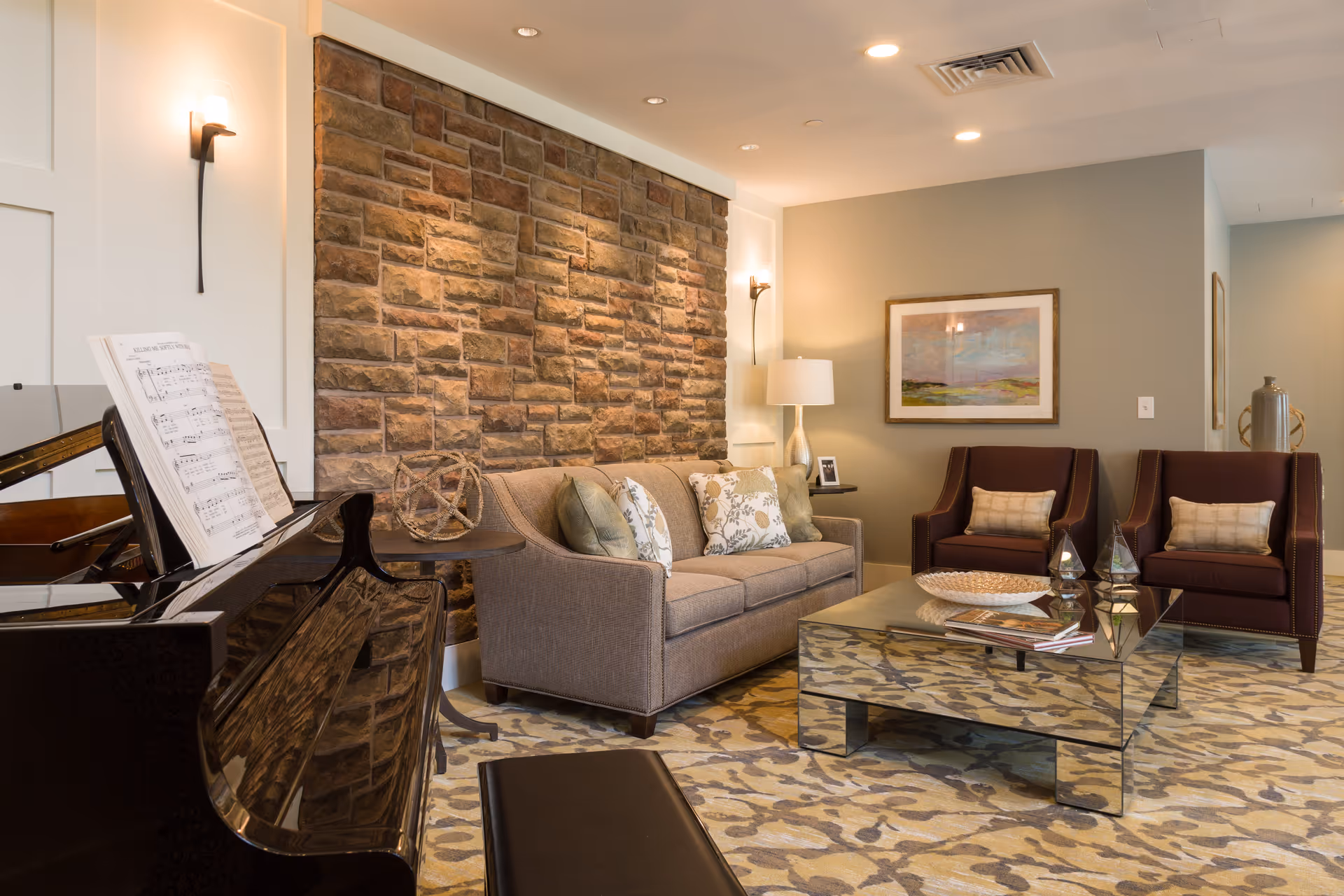 A cozy living room with a stone accent wall, a beige sofa with decorative pillows, two maroon armchairs, a glass coffee table with books and decorative items, a piano with sheet music, a floor lamp, and framed artwork on the wall.