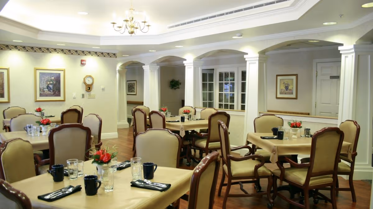 A well-lit dining room with several tables covered in beige tablecloths, each set with black napkins, glassware, and small floral centerpieces. The room features upholstered wooden chairs, framed artwork on the walls, white columns, and a chandelier hanging from the ceiling.