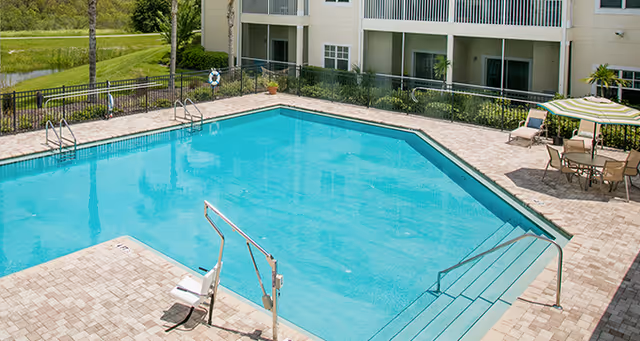 Outdoor swimming pool with clear blue water surrounded by a paved deck with lounge chairs and tables. The pool area is enclosed by a black metal fence and adjacent to a multi-story residential building with balconies. Green grass and trees are visible in the background.
