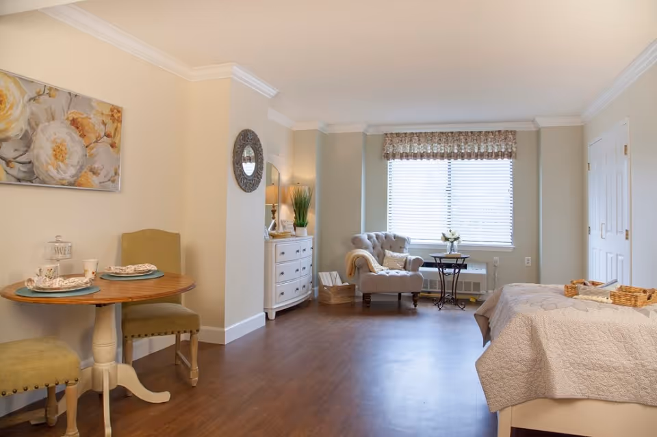 Bright, furnished studio-style bedroom with a bed, tufted armchair by a window, small dining table, dresser, and hardwood floors.