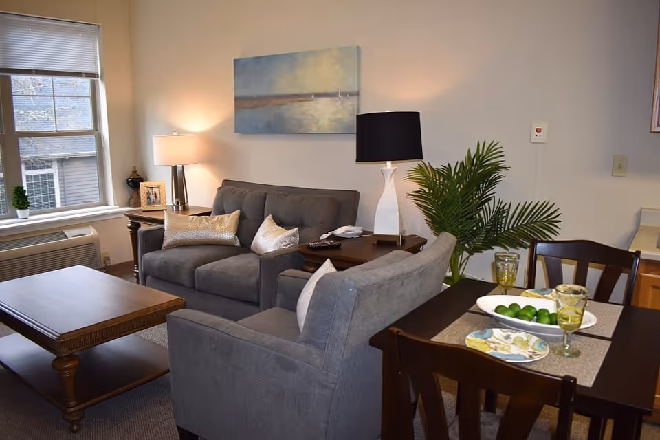 A cozy living room area with a gray loveseat and armchair, a wooden coffee table, and a side table with a lamp and framed photo. There is a window with blinds, a wall painting above the loveseat, and a dining table set with plates, glasses, and a bowl of limes. A green plant is placed next to the dining table.