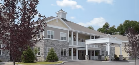 Front exterior of a two-story senior living building with stone and siding facade, a covered entrance/porte-cochere, and surrounding landscaping.