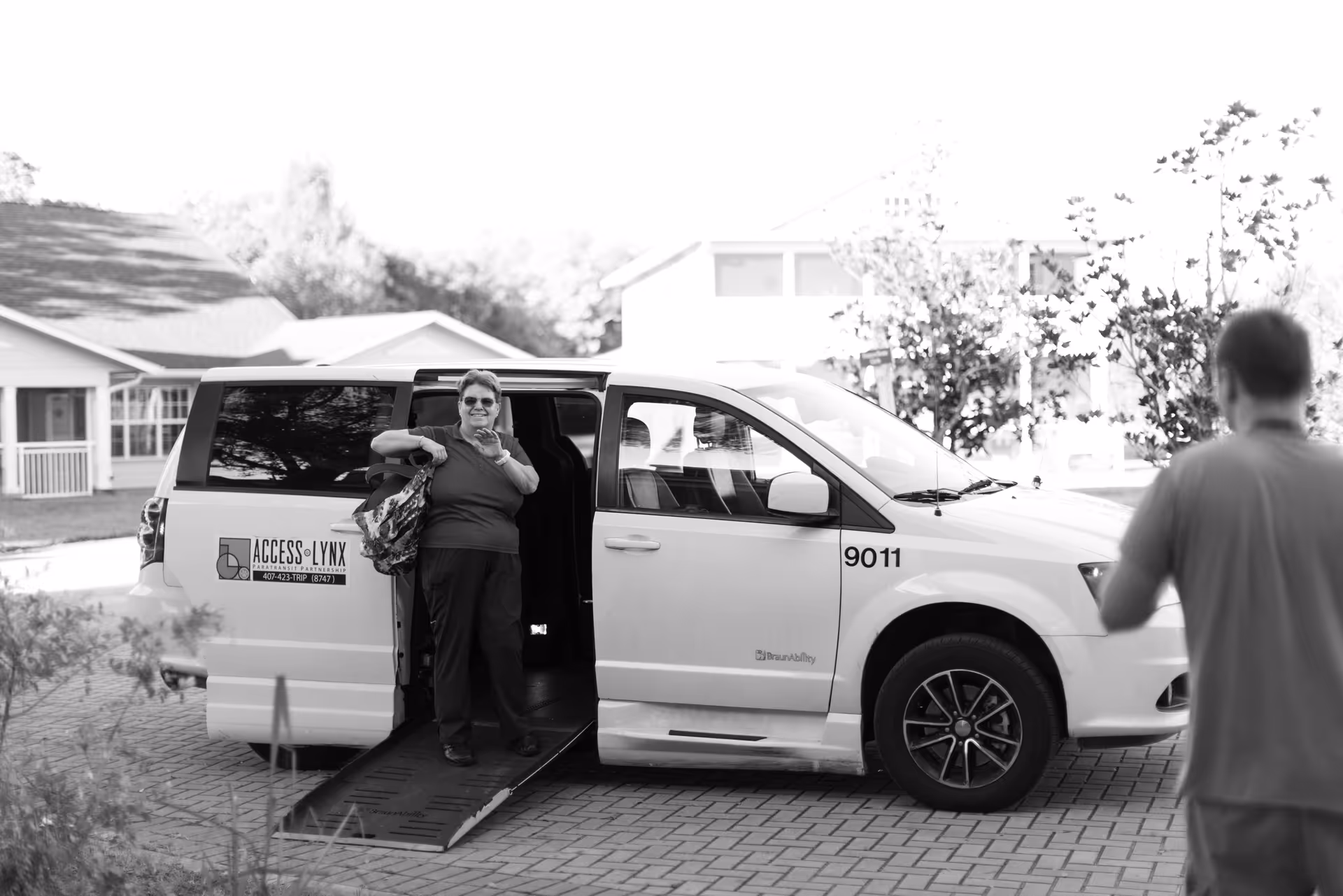 A woman with a bag is stepping out of a white van with a wheelchair accessibility ramp extended. The van has the logo and text 'ACCESS LYNX' on the side. A man stands nearby facing the van. The scene is outdoors in a residential neighborhood with houses and trees in the background.