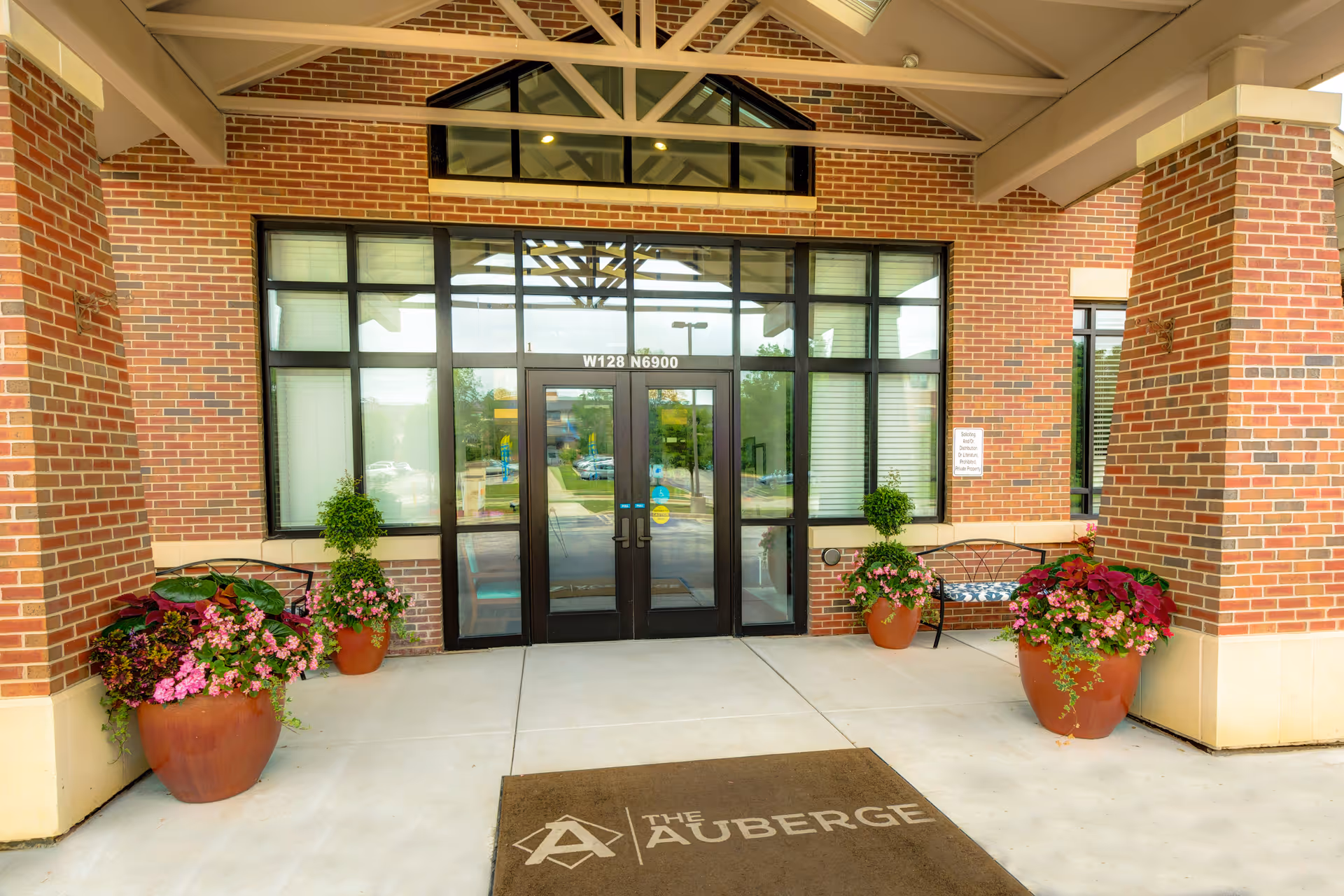 The brick entrance of The Auberge with double glass doors, large windows, potted flowers, benches, and a welcome mat.