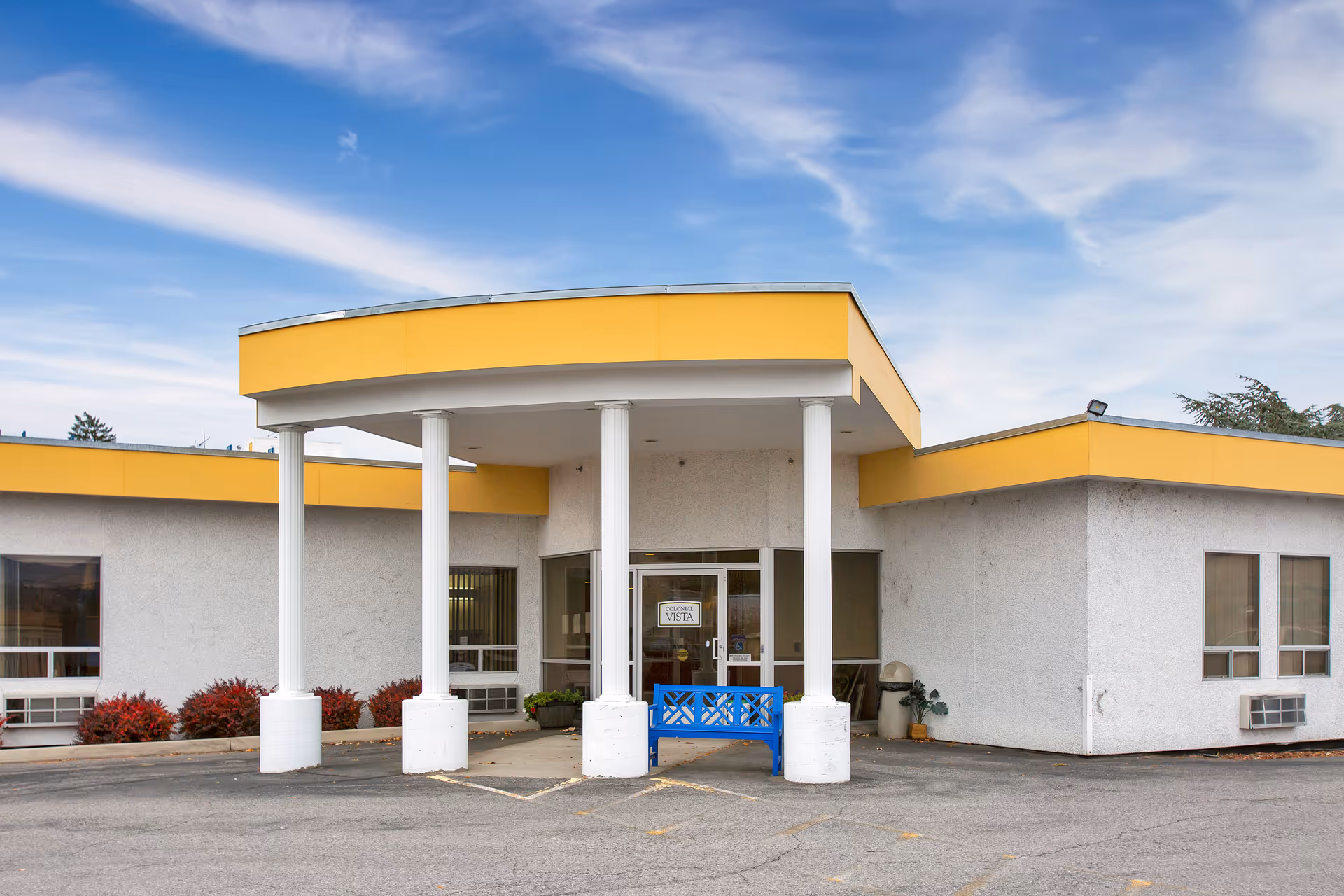 Front exterior view of Colonial Vista Post Acute facility with a yellow and white building, four white columns supporting a covered entrance, a blue bench in front, and a clear sky with some clouds above.