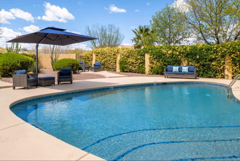 Outdoor swimming pool with clear blue water surrounded by a concrete deck. There are several cushioned wicker chairs and a sofa with blue cushions and pillows, along with a large blue patio umbrella. The area is landscaped with green bushes, trees, and a beige wall in the background under a partly cloudy sky.